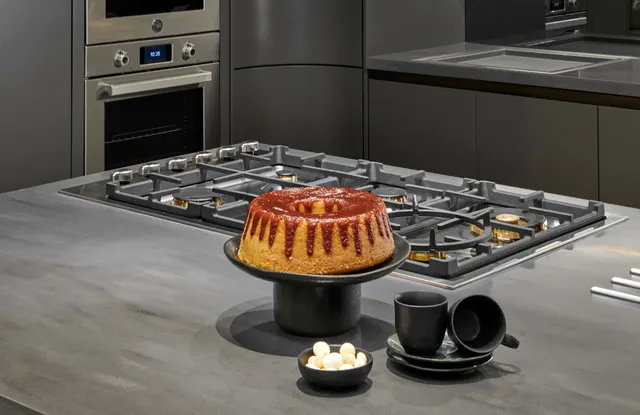 Bolo sobre a bancada de uma ilha com cooktop a gás, com fornos de embutir em aço inox ao fundo