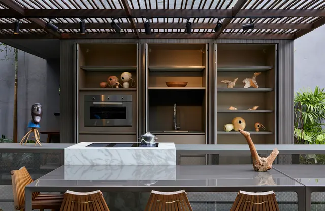 Cozinha gourmet externa sob pérgola, com forno de embutir, torneira em nicho e ilha de mármore com cooktop de indução