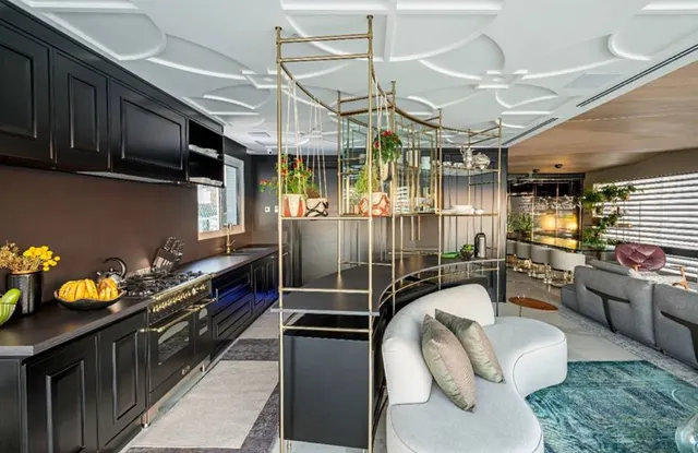 Cozinha preta com fogão de bancada em aço e dourado, estante dourada curva e sala de estar integrada