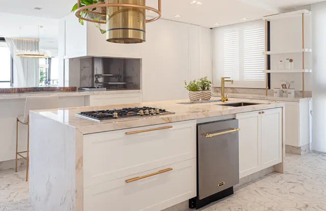 Ilha de mármore com cooktop a gás, torneira dourada e lava-louças em aço inox ao lado de uma coifa cilíndrica dourada