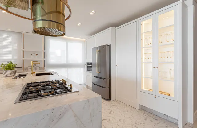 Cozinha branca com geladeira inox, cristaleira iluminada, cooktop na ilha de mármore e coifa cilíndrica dourada